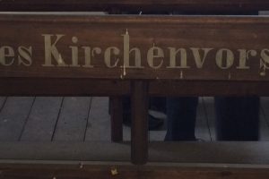 Bank in der Herborner Kirche mit der Aufschrift "Sitze des Kirchenvorstandes", im Hintergrund sind die Beine von drei Personen zu sehen