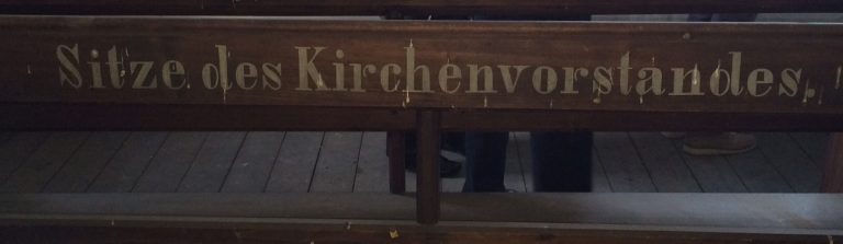 Bank in der Herborner Kirche mit der Aufschrift "Sitze des Kirchenvorstandes", im Hintergrund sind die Beine von drei Personen zu sehen